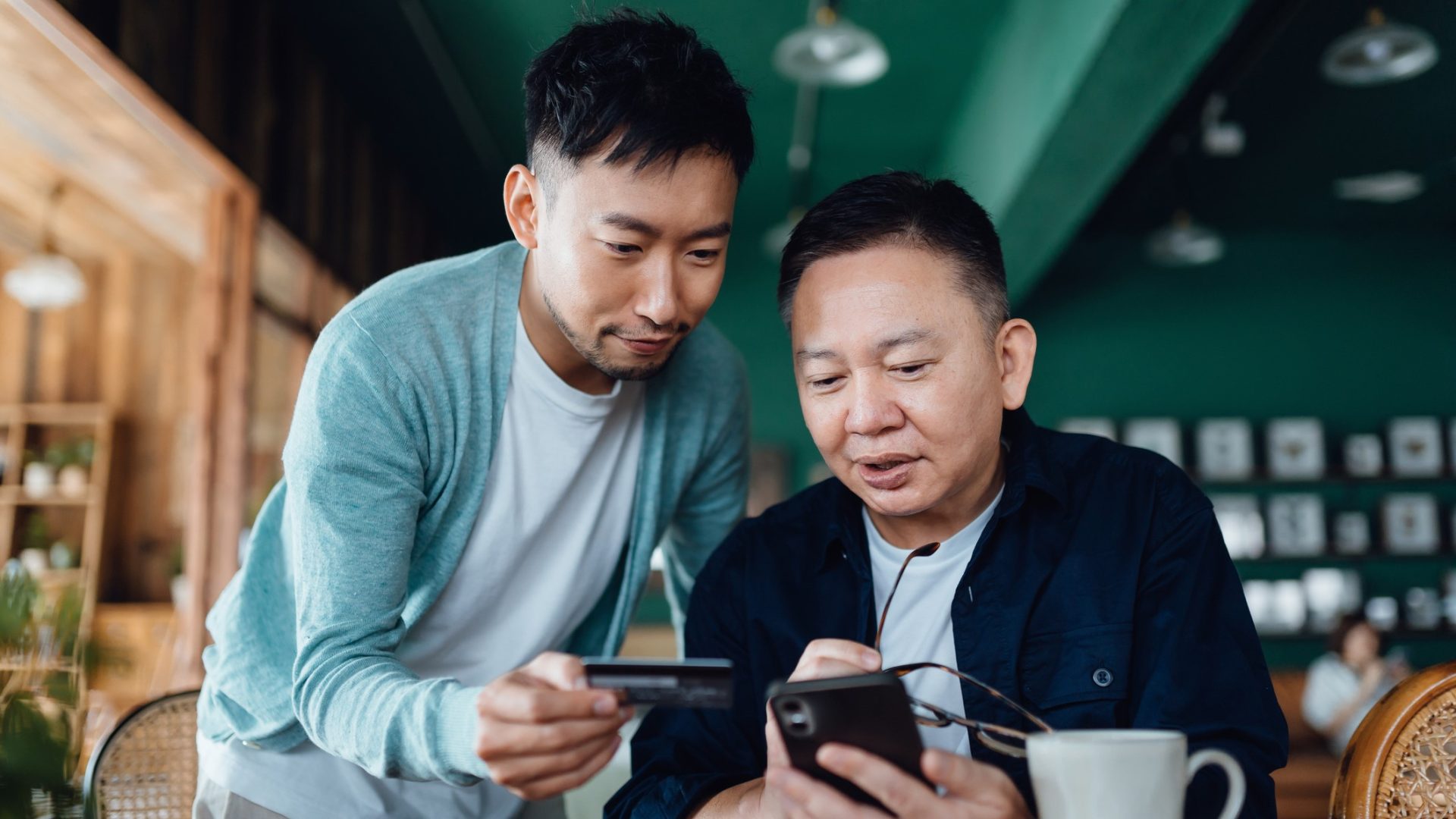 Joyful Asian senior father and son shopping online with mobile app on smartphone together and making payment with credit card online at home.